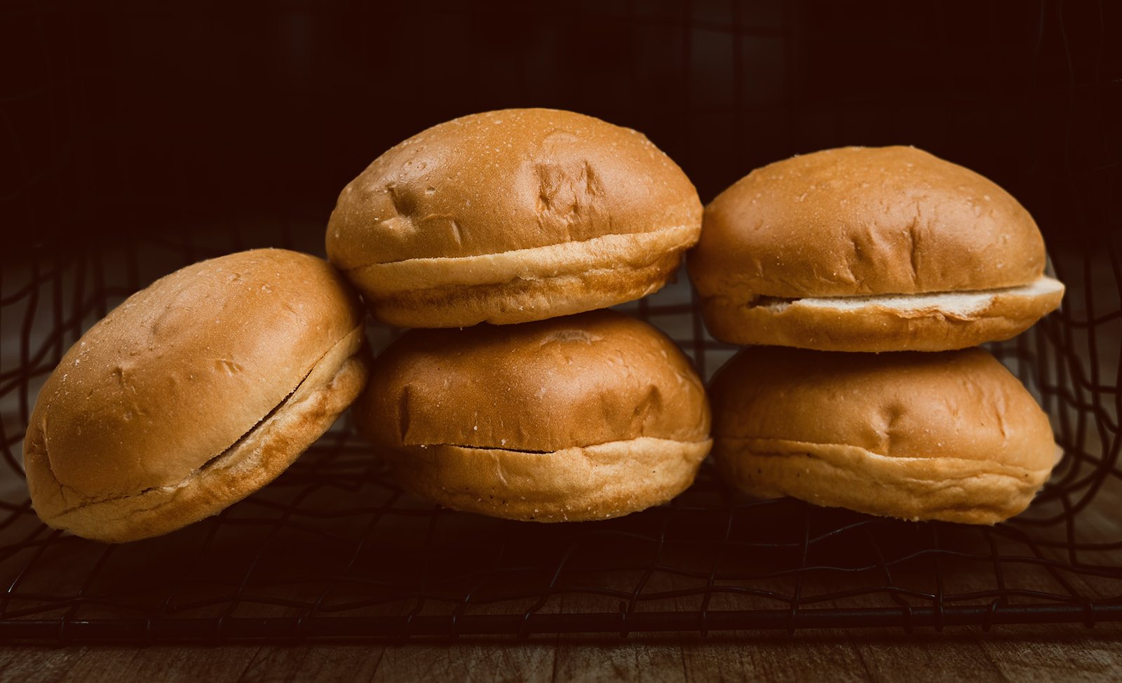 Pre-sliced Coca hamburger buns with pillowy golden crowns on a cooling rack, warm light against a deep shadow background.