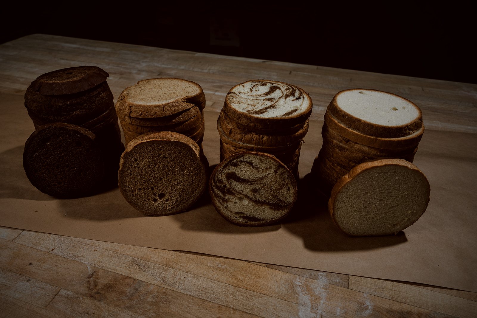 Cross-section slices of pumpernickel, rye, marble, and sourdough deli loaves arranged on a wooden bench against a dark background.
